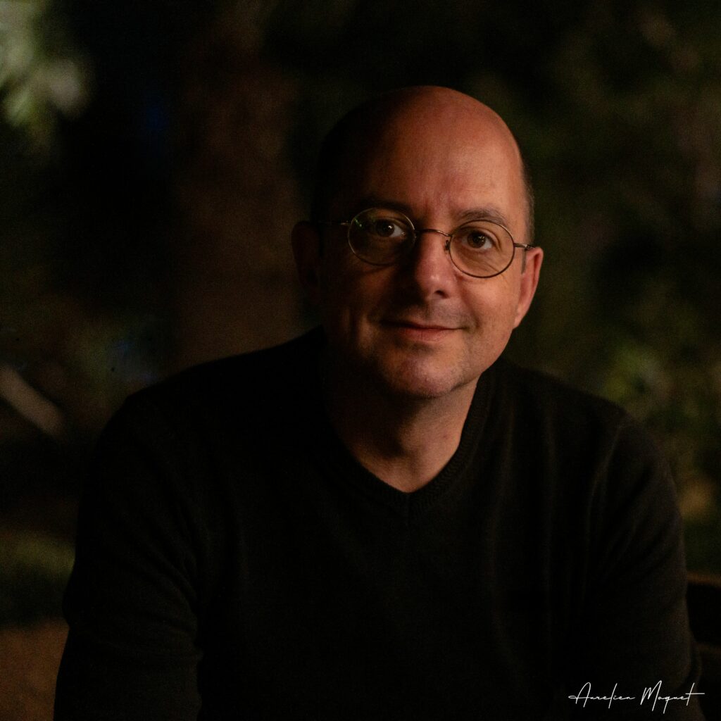 Portrait d'Aurélien Moquet, créateur du site moquet.org, souriant à l'appareil photo dans un jeu de lumière tamisée et un décor de nature en arrière-plan