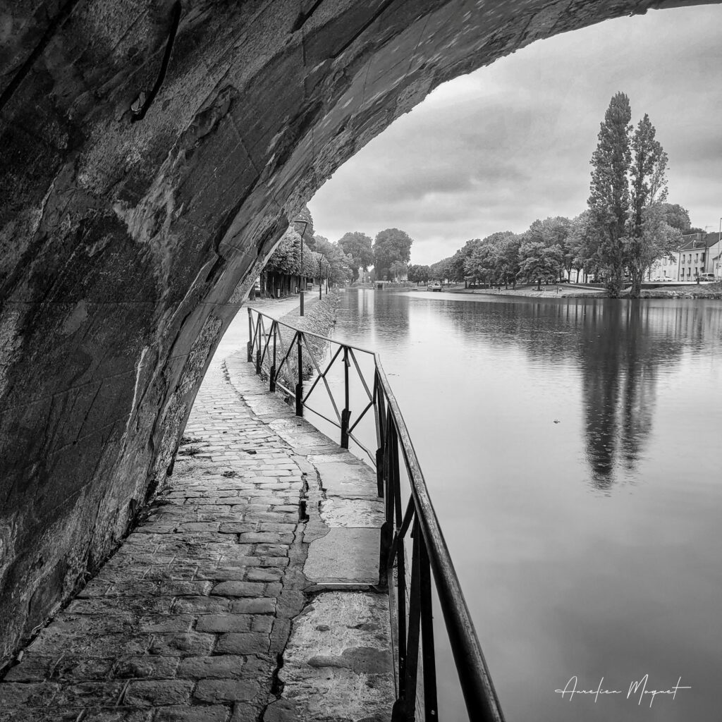 Vue en noir et blanc du canal de Vierzon sous une arche de pont par temps de pluie, Aurélien Moquet